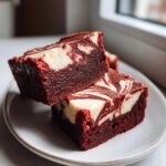 Two rich, fudgy red velvet cheesecake brownies with white cream cheese swirls stacked on a light plate.
