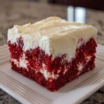 Close-up of a square slice of vibrant red velvet poke cake topped with thick white cream cheese frosting.