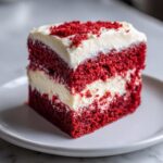 Close-up of a perfectly layered slice of red velvet poke cake topped with thick cream cheese frosting and red crumbs.