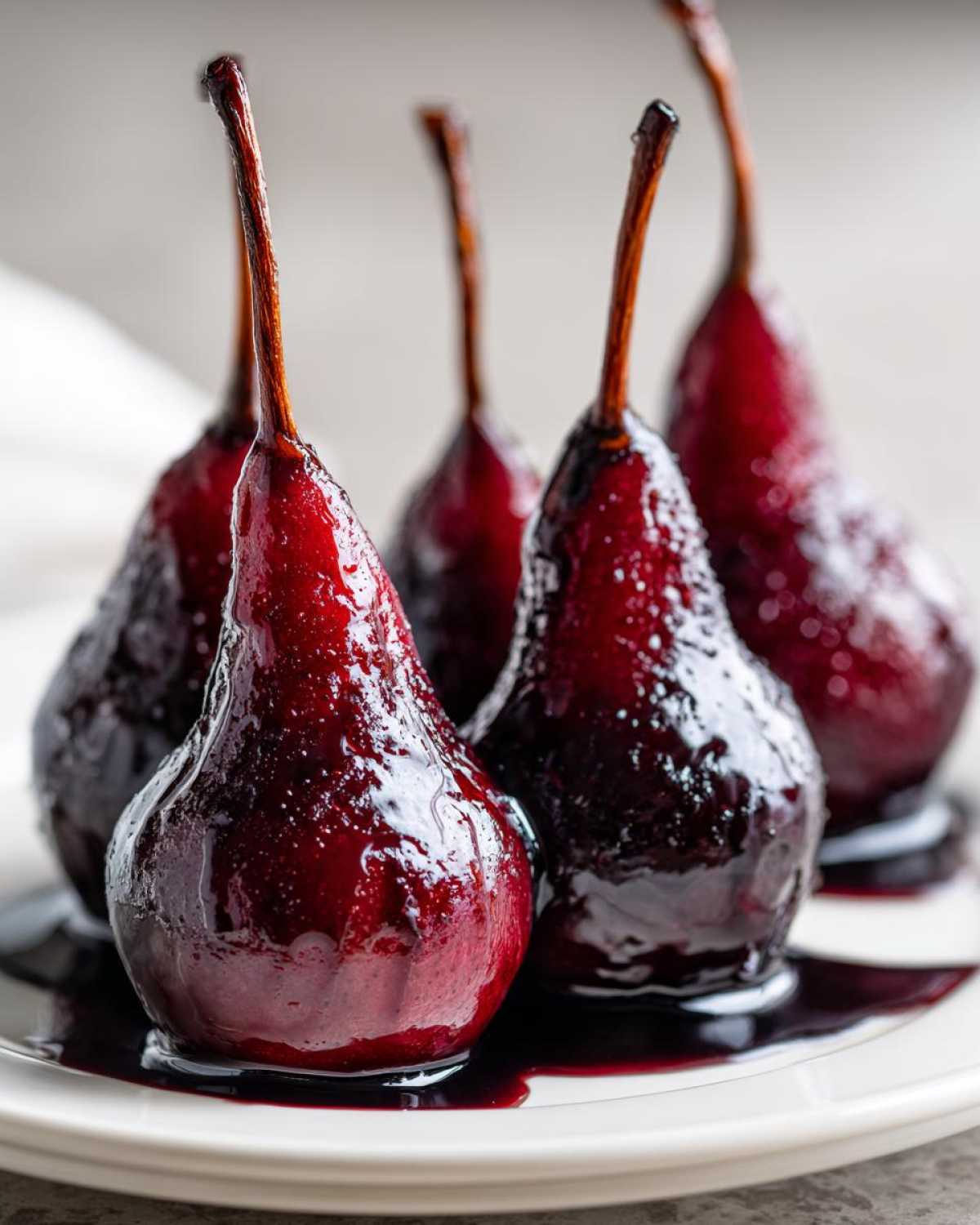 Four glistening, deep ruby red wine poached pears sitting in their syrup on a white plate.