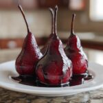 Three glistening, deep red wine poached pears served on a white plate with rich syrup.