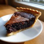 A close-up of a slice of rich chocolate chess pie with a gooey center and flaky crust on a white plate.