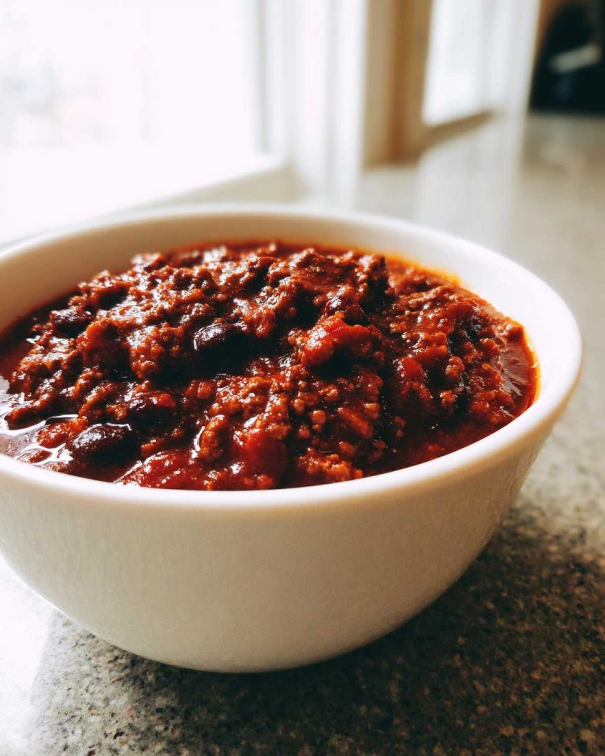 A close-up of a white bowl filled with rich, thick crockpot chili featuring ground meat and beans.