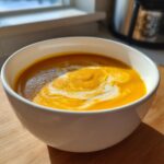 A close-up of vibrant orange roasted butternut squash soup topped with a swirl of cream and black pepper.