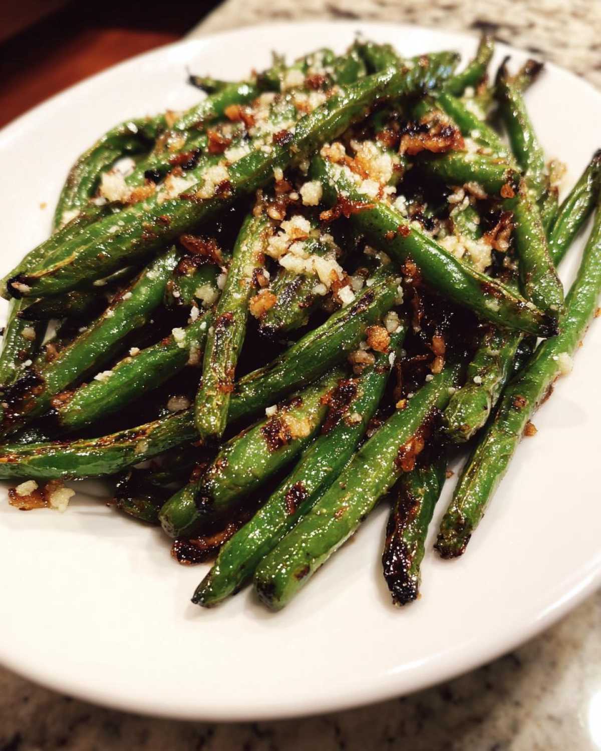 A close-up of roasted, slightly charred green beans topped with minced garlic and seasoning on a white plate.