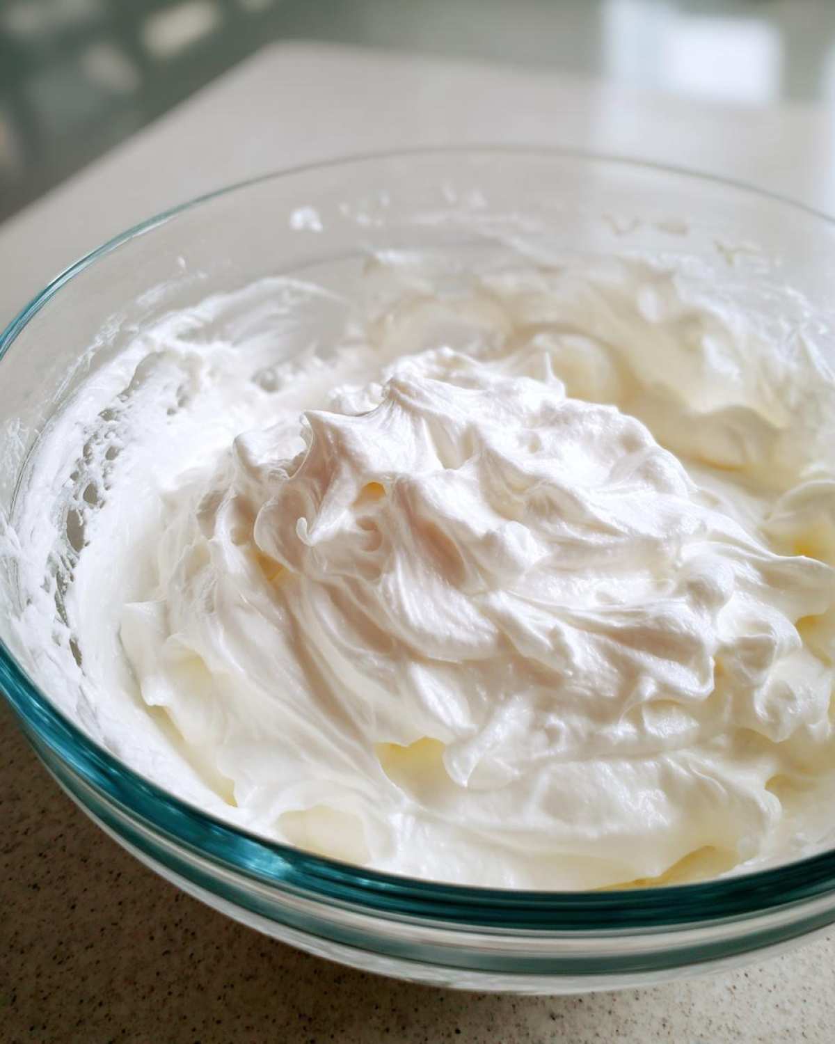 Close-up of thick, glossy white royal icing recipe mixture whipped to stiff peaks in a clear glass bowl.