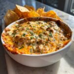 A close-up of a baked sausage cream cheese dip, bubbly and topped with melted cheese and parsley, served with tortilla chips.