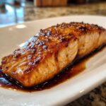 Close-up of a perfectly cooked, glazed salmon fillet resting in a pool of savory sauce on a white plate.