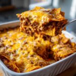A spoonful of cheesy Dorito Casserole, showing layers of ground meat and melted cheese being lifted from a white baking dish.