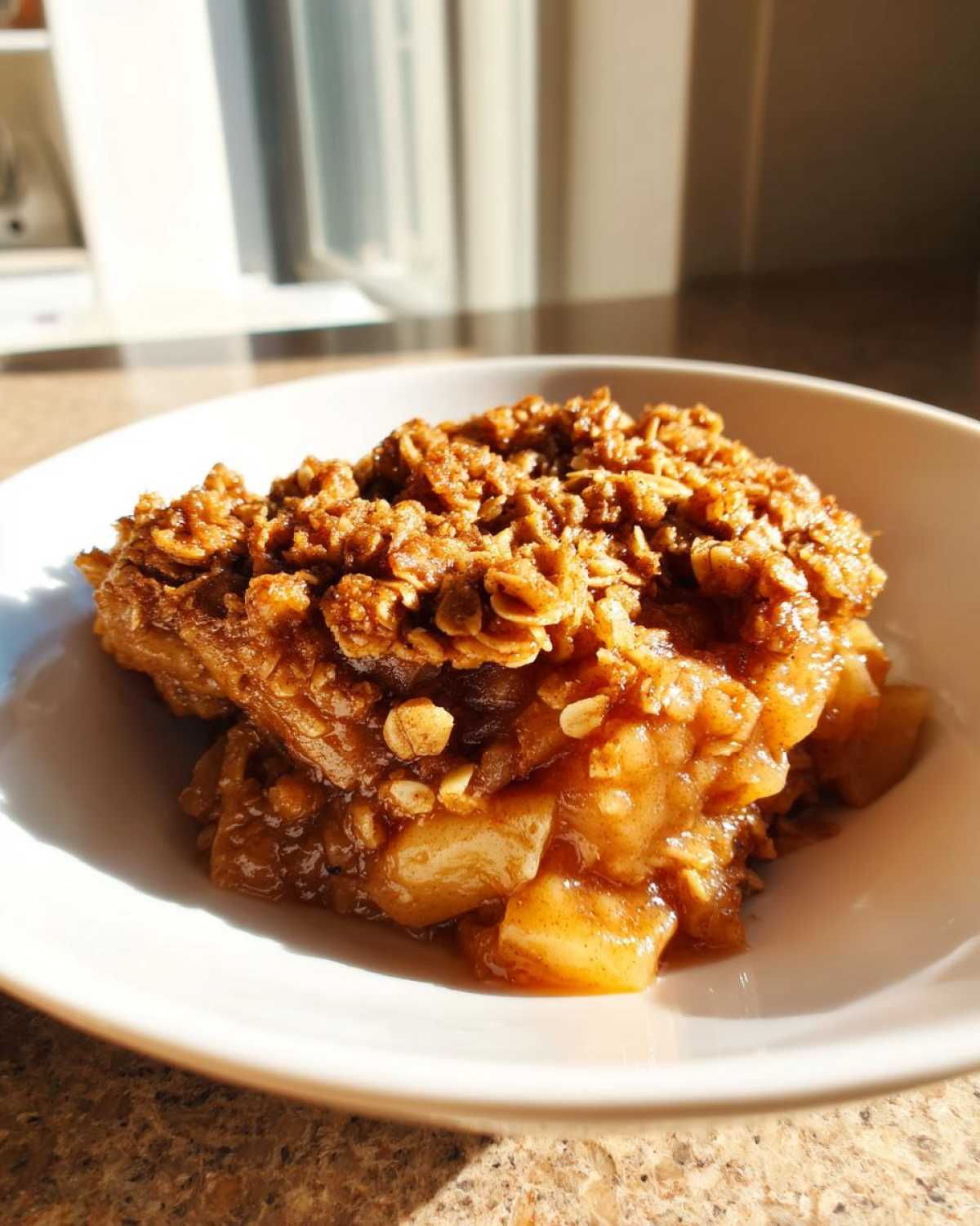 A close-up of a serving of warm apple crisp in a white bowl, featuring soft spiced apples and a crunchy oat topping.