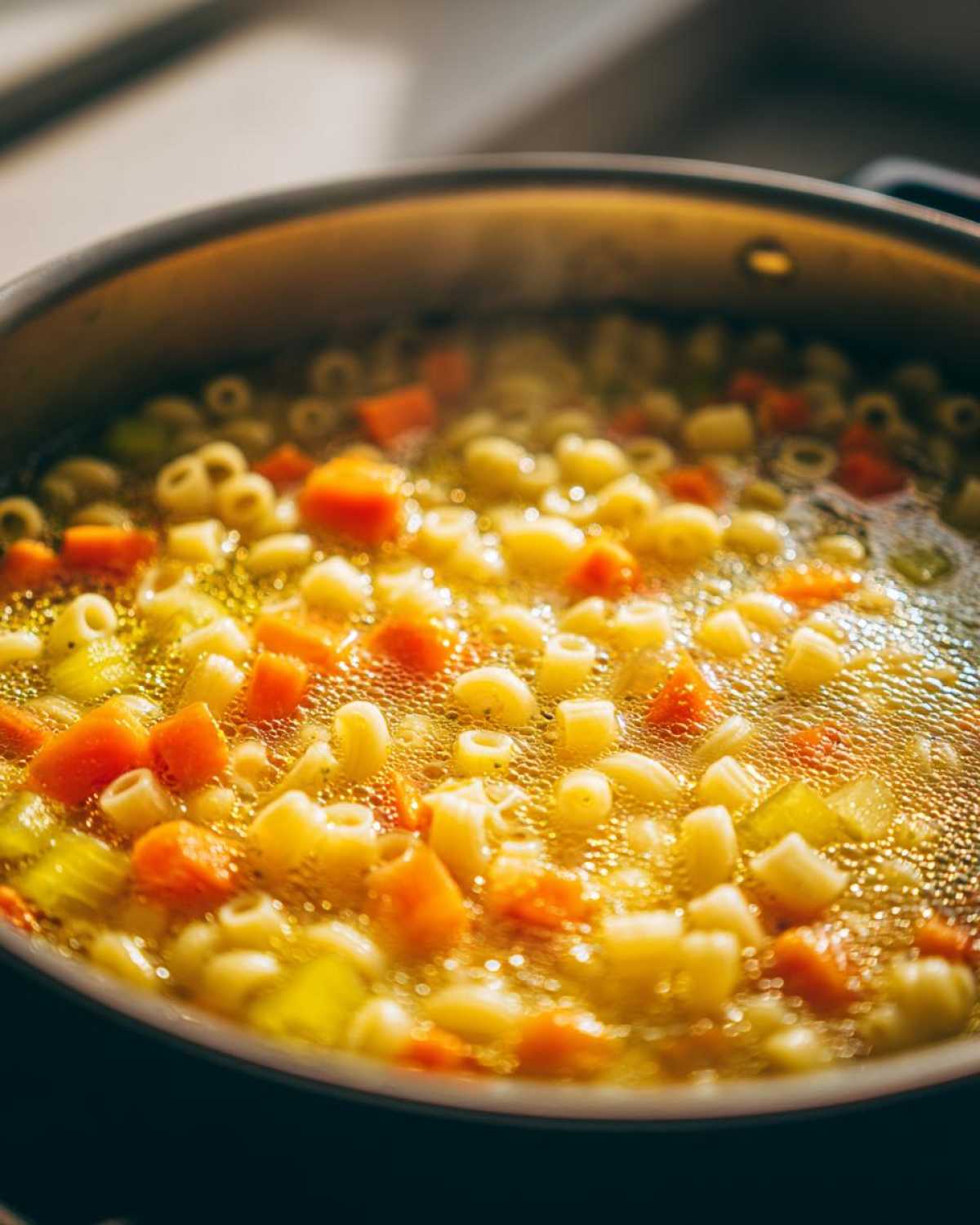 Close-up of italian penicillin soup simmering in a pot, showing small pasta shapes, diced carrots, and celery in a rich broth.