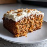 A close-up of a moist slice of carrot cake recipe topped with cream cheese frosting and nuts.