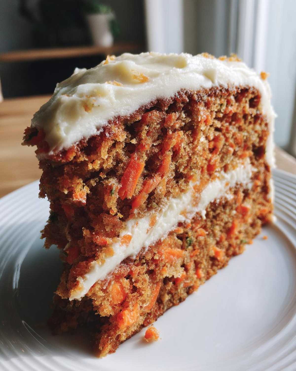 A close-up of a thick slice of carrot cake recipe, showing visible shredded carrots and cream cheese frosting layers.
