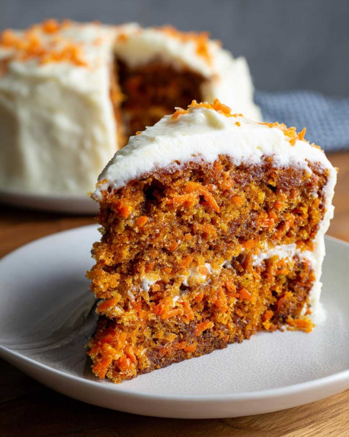 A close-up of a thick slice of moist carrot cake recipe with visible shredded carrots and cream cheese frosting.