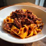 Close-up of tender shredded beef ragu generously coating wide pappardelle pasta in a white bowl.