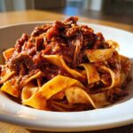 A close-up of rich, shredded beef ragu generously coating wide pappardelle pasta in a white bowl.
