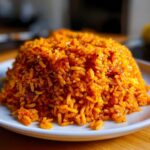 Close-up of a mound of vibrant orange Nigerian jollof rice served on a white plate.