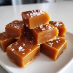 A stack of soft, chewy caramels sprinkled with coarse sea salt on a white plate.