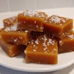 A stack of soft, chewy caramels sprinkled generously with flaky sea salt on a white plate.