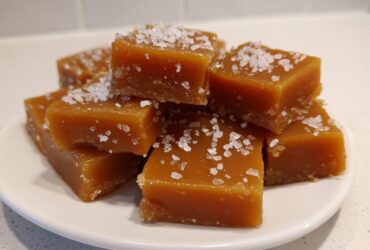 A stack of soft, chewy caramels sprinkled generously with flaky sea salt on a white plate.