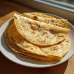 A stack of freshly made, soft corn tortillas with light brown char marks, sitting on a white plate.