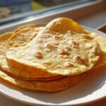 A stack of freshly made, soft corn tortillas with light brown toasted spots, resting on a white plate near a sunlit window.