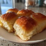 Four golden brown, soft dinner rolls brushed with a shiny glaze, served on a white plate.