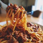A fork lifting spaghetti noodles coated in rich, meaty bolognese sauce from a white plate.