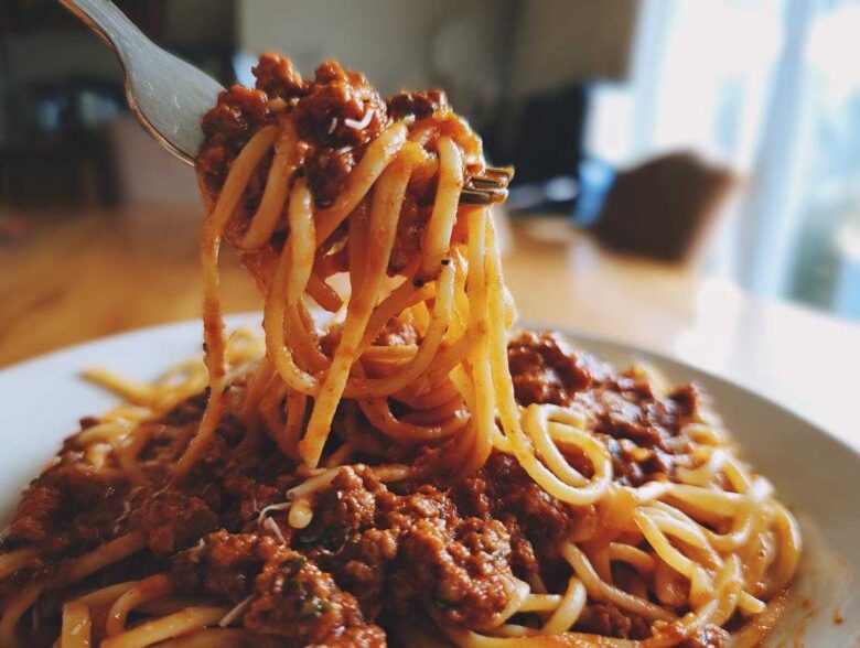 A fork lifting spaghetti noodles coated in rich, meaty bolognese sauce from a white plate.