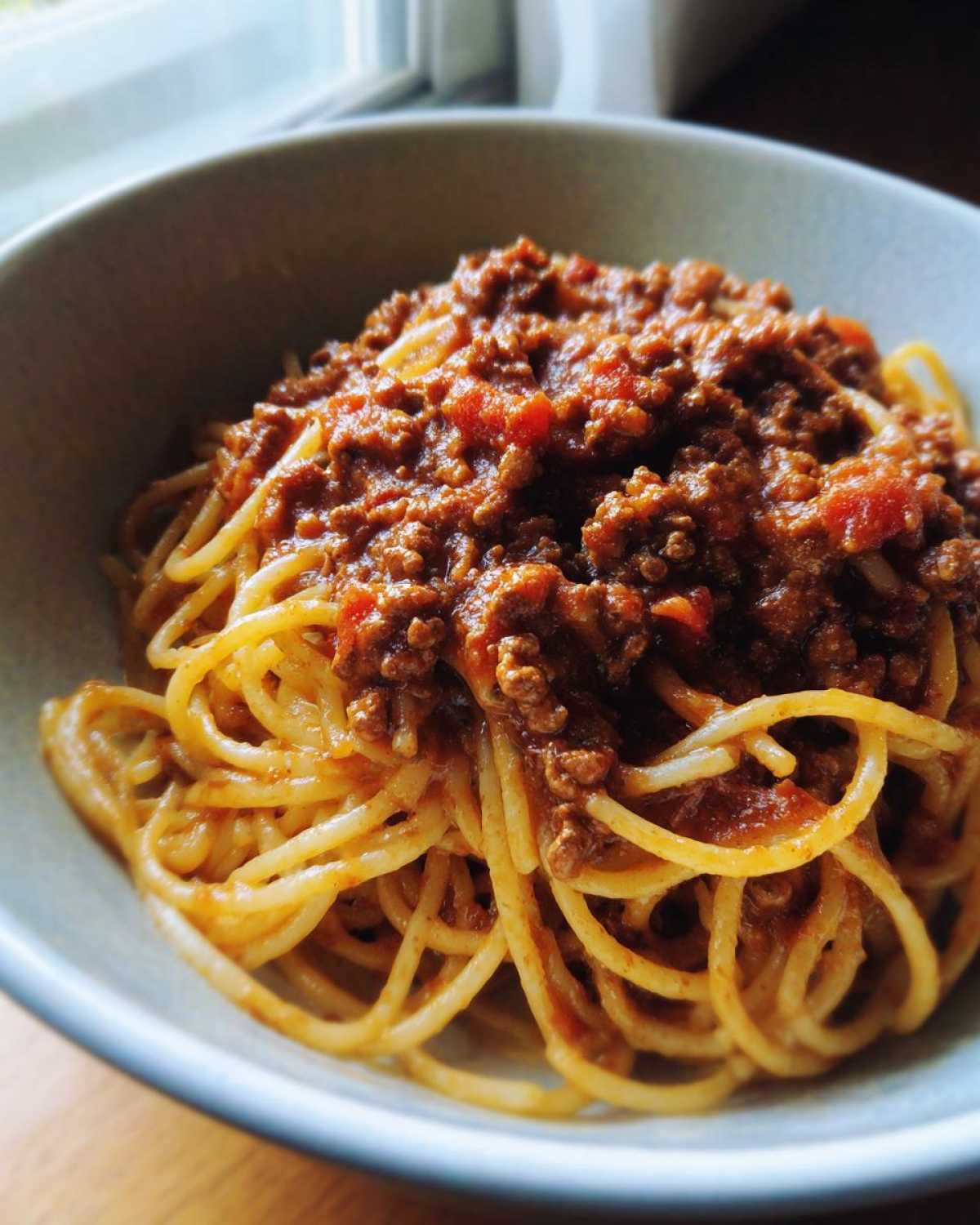 A close-up of spaghetti noodles generously topped with rich, meaty bolognese sauce in a light-colored bowl.