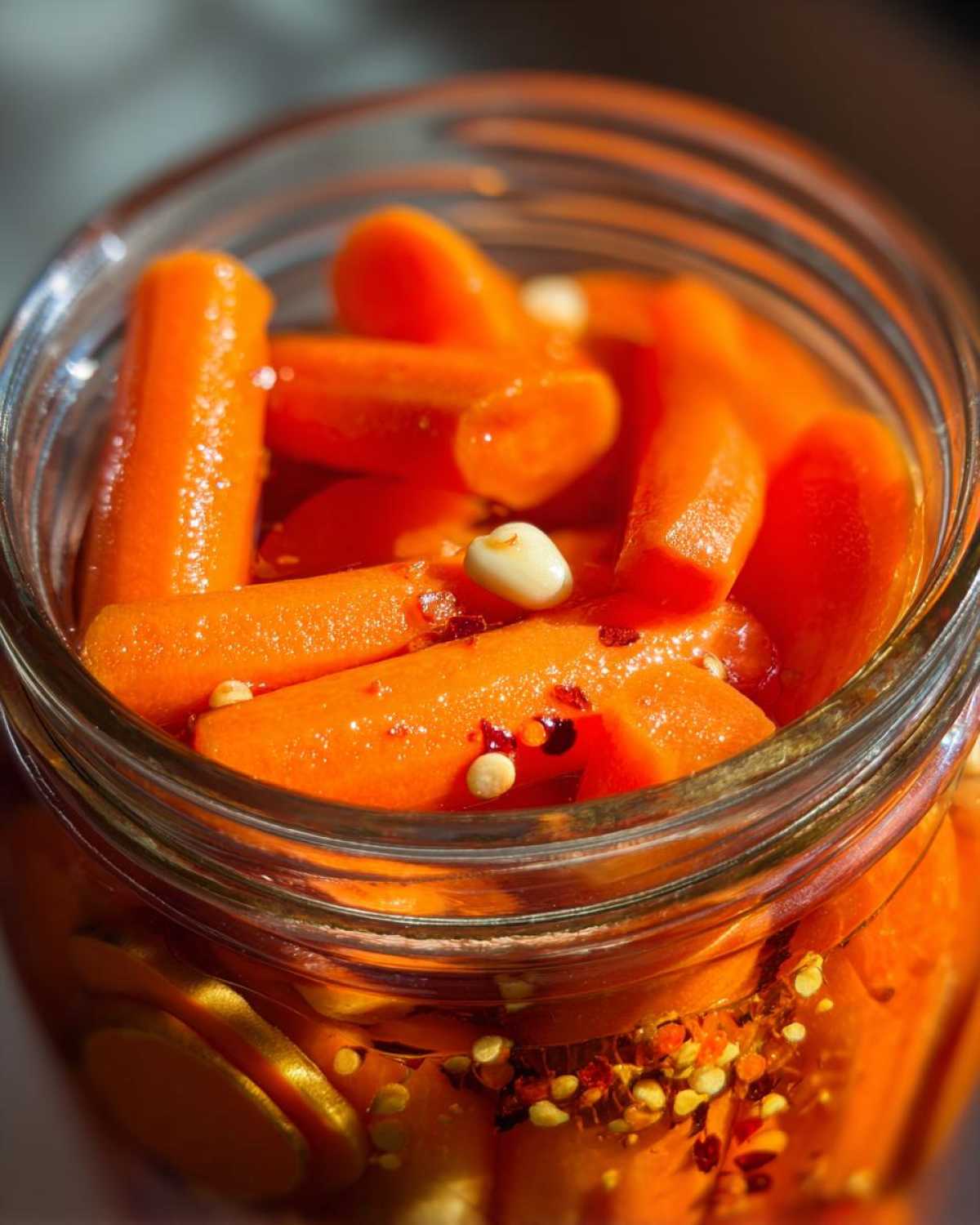 Close-up of bright orange pickled carrots submerged in brine with garlic cloves and red pepper flakes in a glass jar.