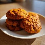 A close-up of a stack of four golden brown, textured persimmon cookies resting on a white plate in bright sunlight.