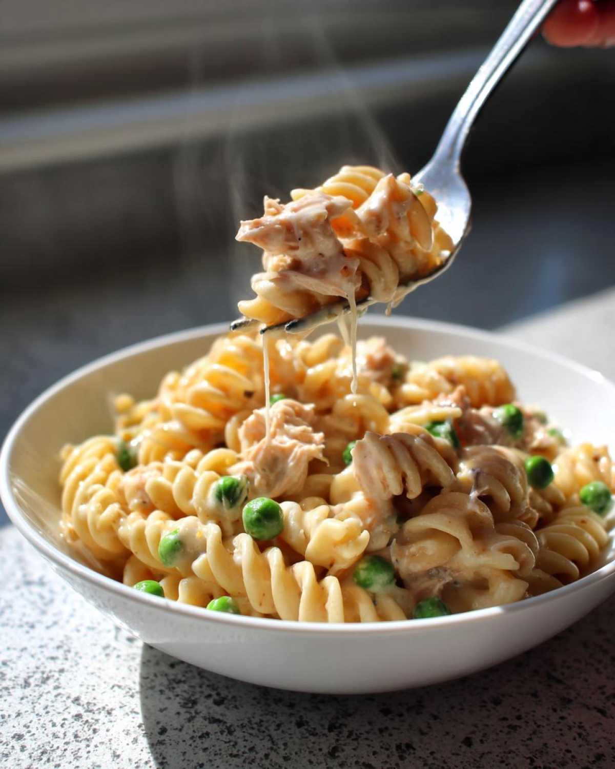 A fork lifts a bite of hot, creamy tuna pasta with fusilli noodles and green peas from a white bowl.