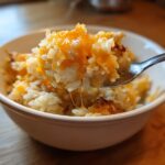 A spoonful of creamy, stovetop cheesy rice topped with melted orange cheddar cheese being lifted from a white bowl.