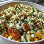 A close-up of a street corn chicken rice bowl featuring seasoned chicken, corn, cotija cheese, and a drizzle of green sauce.