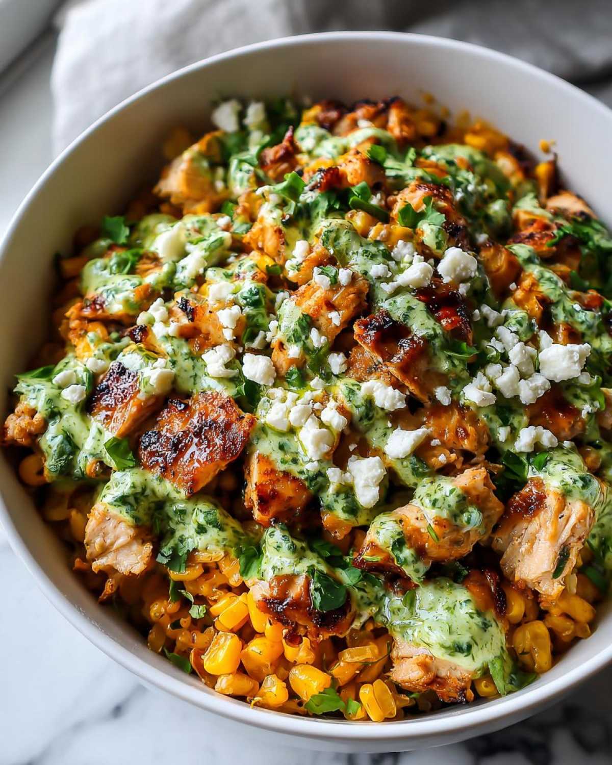Close-up of a street corn chicken rice bowl topped with creamy green sauce and crumbled white cheese.