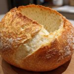 A golden-brown, round loaf of artisan bread with a scored top, ready to be turned into sturdy bread bowls.