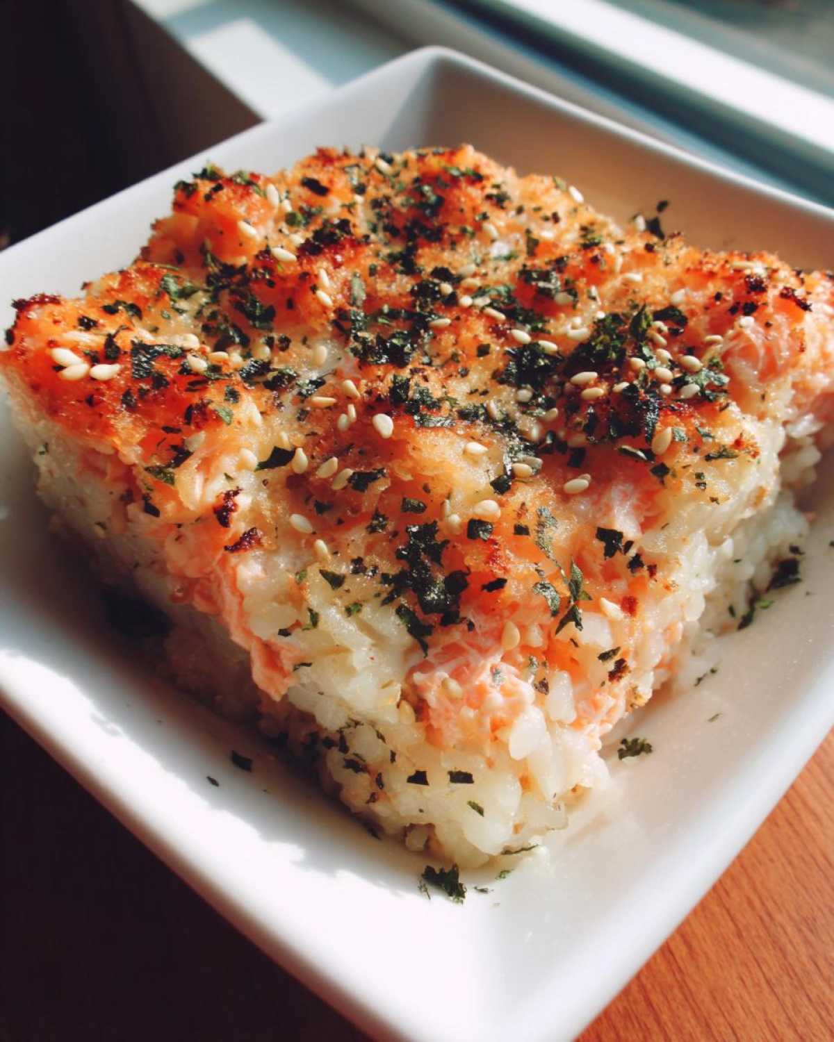 A square serving of baked salmon sushi bake, topped with toasted sesame seeds and furikake seasoning.