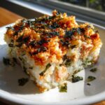 A square slice of baked sushi bake showing layers of rice, seafood, and a crispy, seasoned topping with nori flakes.