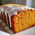 Close-up of a moist, orange-hued sweet potato pound cake topped with thick white icing and orange zest.