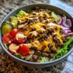 A close-up of a hearty cheeseburger bowl featuring seasoned ground beef, melted cheddar cheese, lettuce, tomatoes, pickles, and a drizzle of special sauce.