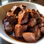 Close-up of tender crockpot steak bites coated in a thick, dark, savory sauce served in a white bowl.