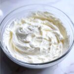 Close-up of thick, stiff white royal icing recipe mixture in a clear glass mixing bowl.