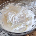 Close-up of thick, perfectly whipped white royal icing recipe mixture in a clear glass bowl.
