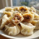 A plate piled high with freshly steamed Traditional Chinese Dumplings, showing the savory meat filling in the cut ones.