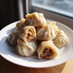 A plate piled high with freshly steamed Traditional Chinese Dumplings, one cut open to show the savory meat filling.