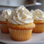 Three moist vanilla cupcakes topped with high swirls of creamy white frosting sitting on a white plate.