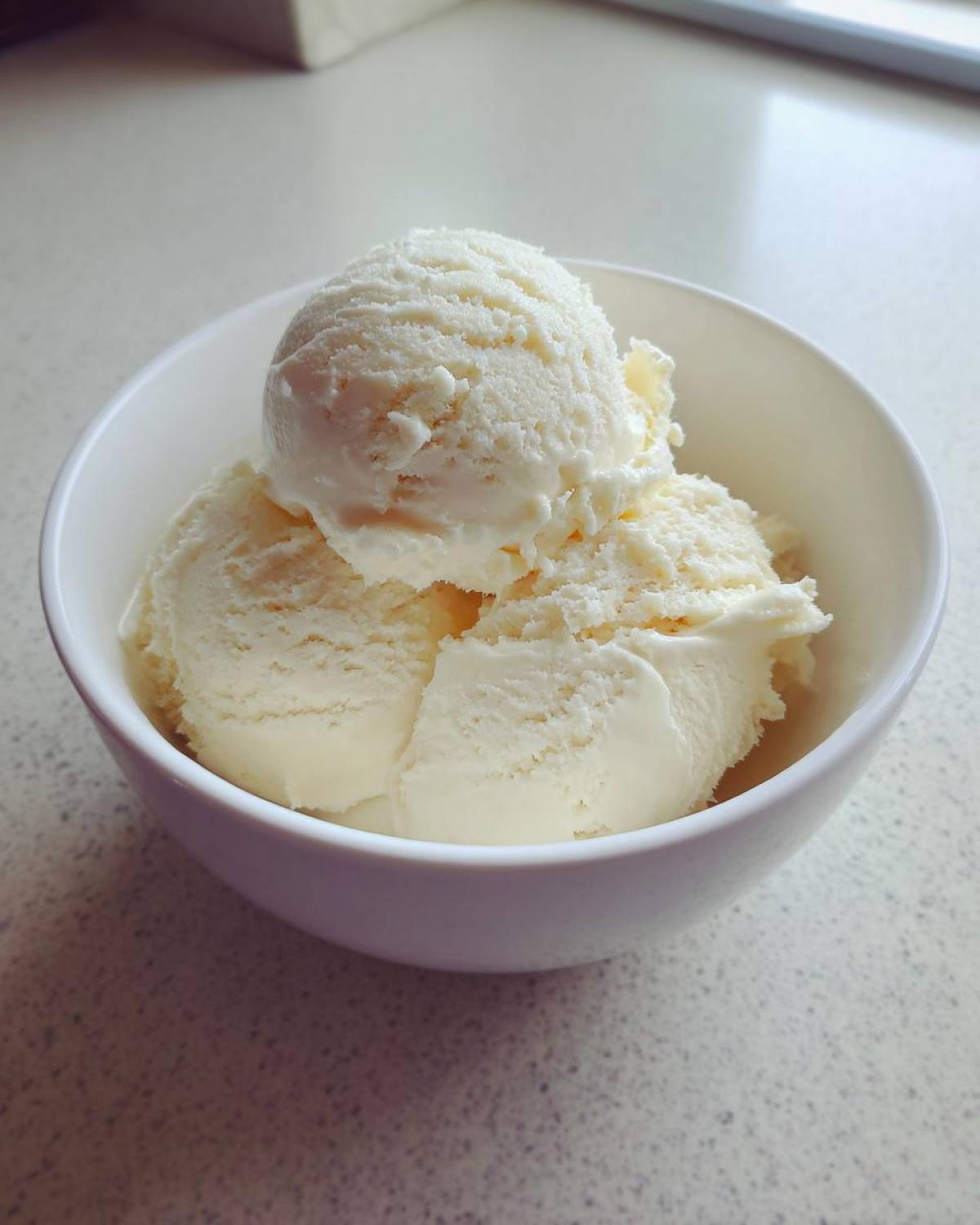 Three scoops of creamy vanilla protein ice cream served in a small white bowl on a light countertop.