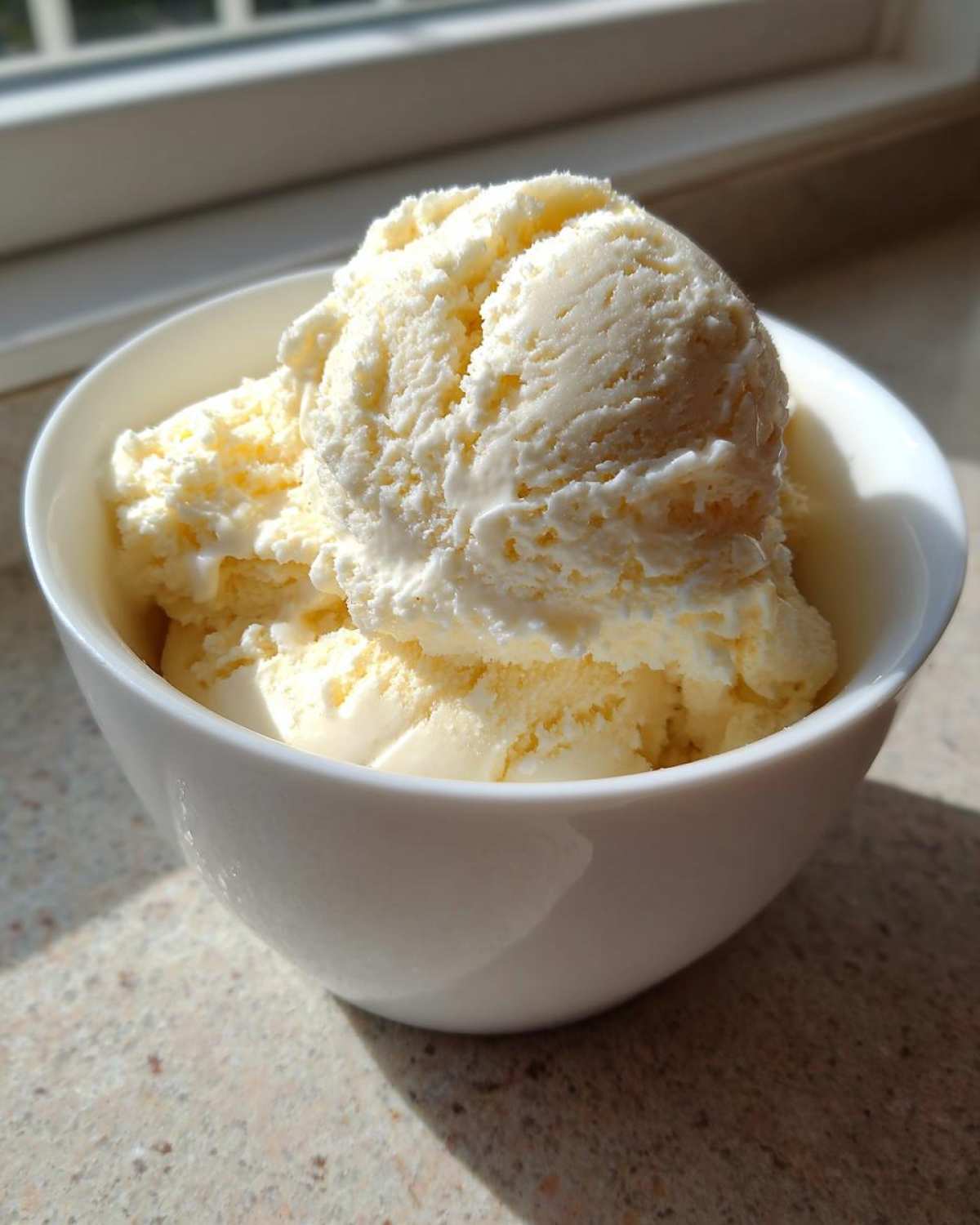 Two scoops of creamy vanilla protein ice cream served in a small white bowl, illuminated by sunlight.