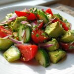 Close-up of a fresh avocado salad featuring chunks of avocado, tomato, cucumber, and red onion on a white plate.